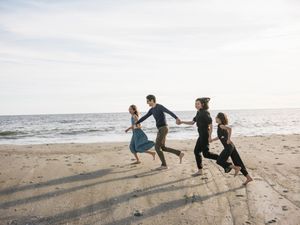 Four friends holding hands and running joyfully along a sandy beach with ocean waves in the background.