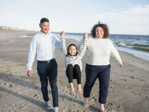 Family of three joyfully swinging their daughter on a sunny beach, with ocean waves and sandy shore in the background.