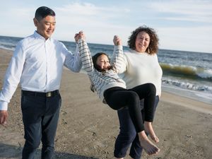 Family of three joyfully swinging child on sunny beach, parents holding hands with laughing girl by ocean waves.