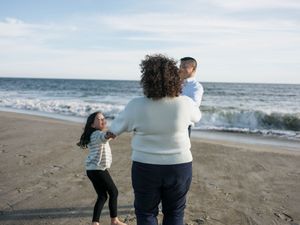 A mother holds her son while her daughter dances on a sandy beach with ocean waves in the background.