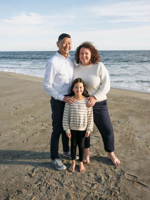 A family of three smiles together on a sandy beach, with ocean waves in the background on a sunny day.