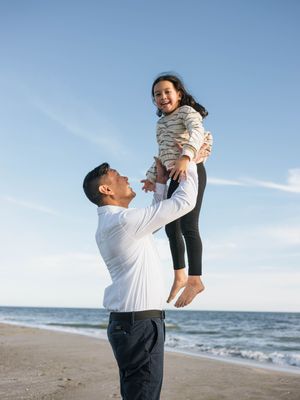 A father joyfully lifts his laughing daughter high in the air on a sunny beach with blue skies.
