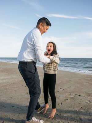 A father and young daughter laugh and play together on a sandy beach with ocean waves in the background.