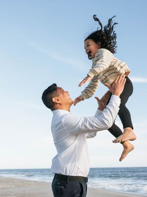 Father joyfully tosses laughing child into the air on a sunny beach with ocean waves in the background.