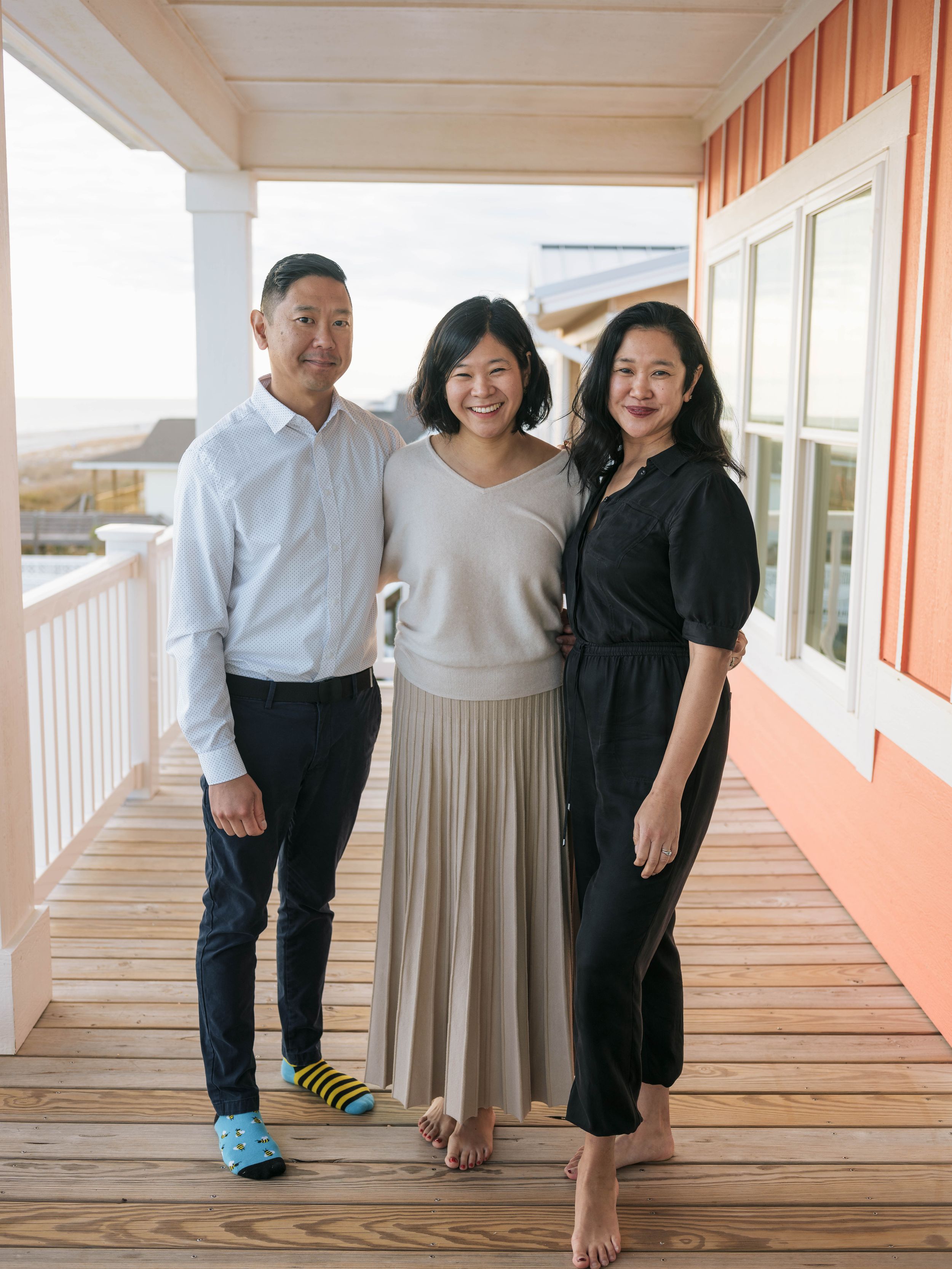Three people smiling together on a wooden porch of a colorful beach house on a sunny day.