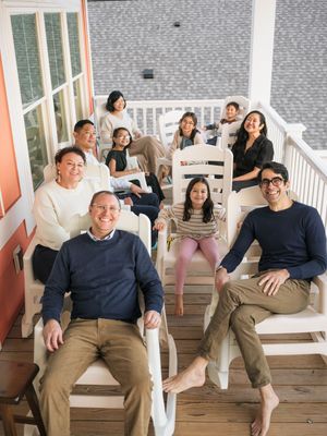 Large family group smiling together on a porch, posing for a portrait in casual attire on a sunny day.