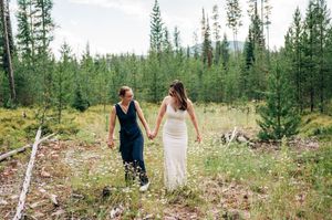 Two brides holding hands walking through a wildflower meadow surrounded by pine trees.