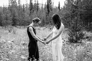 Two women hold hands in a wildflower meadow with forest backdrop in a black and white wedding photo.