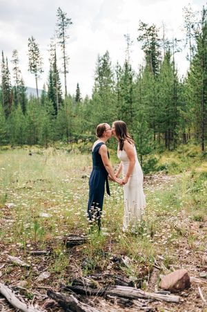 Two brides share a kiss in a wildflower meadow surrounded by evergreen forest on their wedding day.