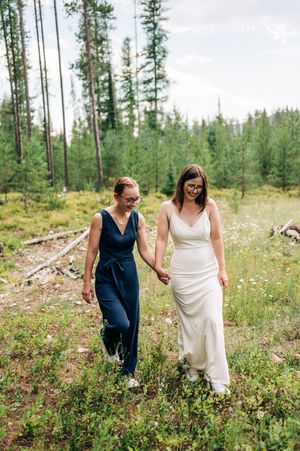 Two brides walk hand-in-hand through a wildflower meadow, one in white gown and one in navy jumpsuit.