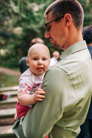 A man in a green shirt tenderly holds a curious baby at an outdoor wedding.