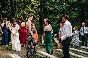 Wedding guests in formal attire mingle outdoors among trees, wearing elegant dresses in white, red, floral, and green.