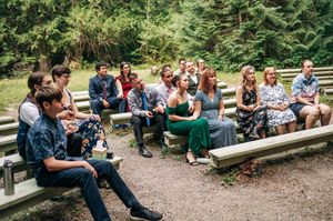 Guests seated on wooden benches at an outdoor wedding ceremony surrounded by lush green forest trees.