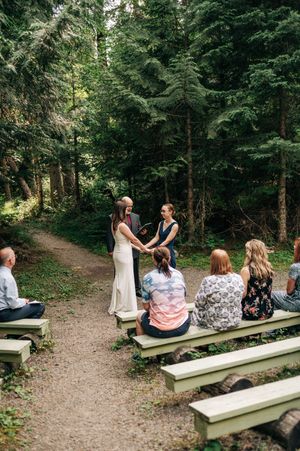 Intimate outdoor wedding ceremony in a forest with guests seated on wooden benches as couple exchanges vows.