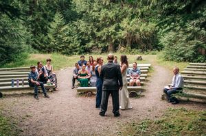 Outdoor wedding ceremony in a forest clearing with guests seated on wooden benches surrounding the couple.