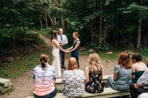 Outdoor forest wedding ceremony with couple exchanging vows before officiant, guests seated on wooden benches among tall green trees.