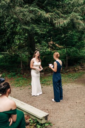 Bride in white gown laughing with bridesmaid in navy dress during outdoor forest wedding ceremony.