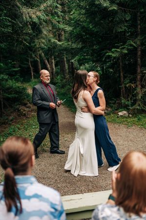 Two brides share a kiss during an intimate outdoor forest wedding ceremony with an officiant nearby.