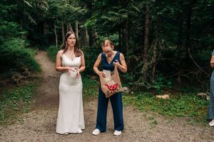 Two women stand on a forest path, one in a white gown and one in a navy jumpsuit holding a gift bag.