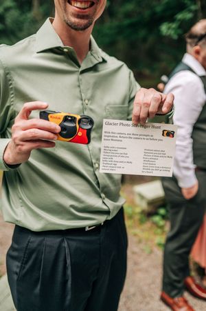 Man holding a Kodak disposable camera and a Glacier Photo Scavenger Hunt card at an outdoor wedding event.