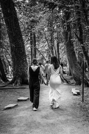 Two women in black and white dresses walk hand-in-hand down a tree-lined forest path at a wedding.