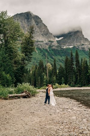Two brides embrace on a rocky riverbank with misty mountains behind them.