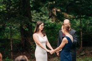 Two women hold hands during an outdoor forest wedding ceremony with an officiant in background.