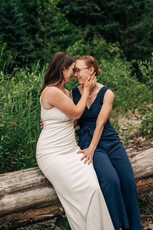 Two brides share a romantic moment sitting on a log, one in white gown and one in navy jumpsuit, surrounded by lush greenery.