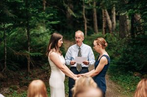 Officiant reads vows as two women hold hands during an intimate outdoor forest wedding ceremony.
