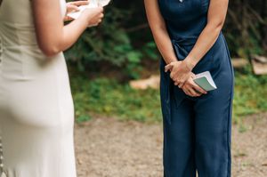 Two women at an outdoor wedding ceremony, one in white dress, other in navy jumpsuit holding small book.