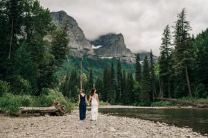 Two brides hold hands by a mountain river surrounded by evergreen trees and misty peaks in Glacier National Park.