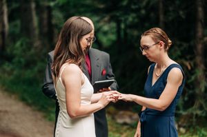 Two brides exchange rings during an intimate outdoor wedding ceremony in a forest setting.