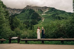 Two brides stand on a wooden fence rail with a dramatic mountain backdrop during their outdoor wedding.