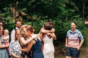 Bride in white dress hugs wedding guest outdoors surrounded by smiling friends and family in a forest setting.