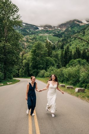 Two brides walk hand-in-hand down a mountain road with lush green scenery.