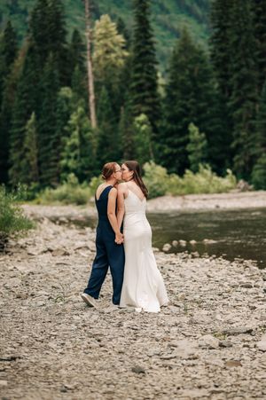 Two brides share a kiss by a rocky riverbank surrounded by lush green pine trees on their wedding day.