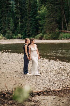 Two brides hold hands on a rocky riverbank, one in a white gown and one in a navy jumpsuit.