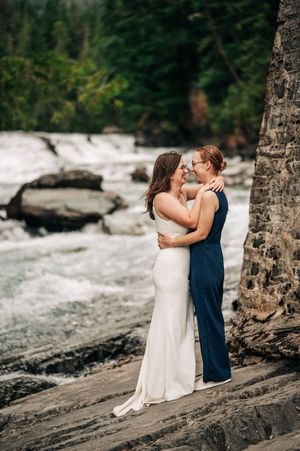 Two brides embrace near a rushing waterfall in a forest setting.