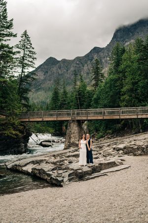 Couple poses on rocky riverbank beneath wooden bridge surrounded by evergreen trees and misty mountains.