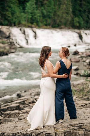 Two brides stand together on rocks in front of a scenic waterfall.
