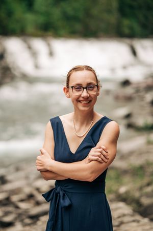 Smiling woman in navy dress with pearl necklace and glasses stands confidently before a waterfall with arms crossed.