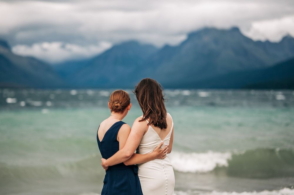 Two women embrace while gazing at a stunning mountain lake under dramatic cloudy skies.
