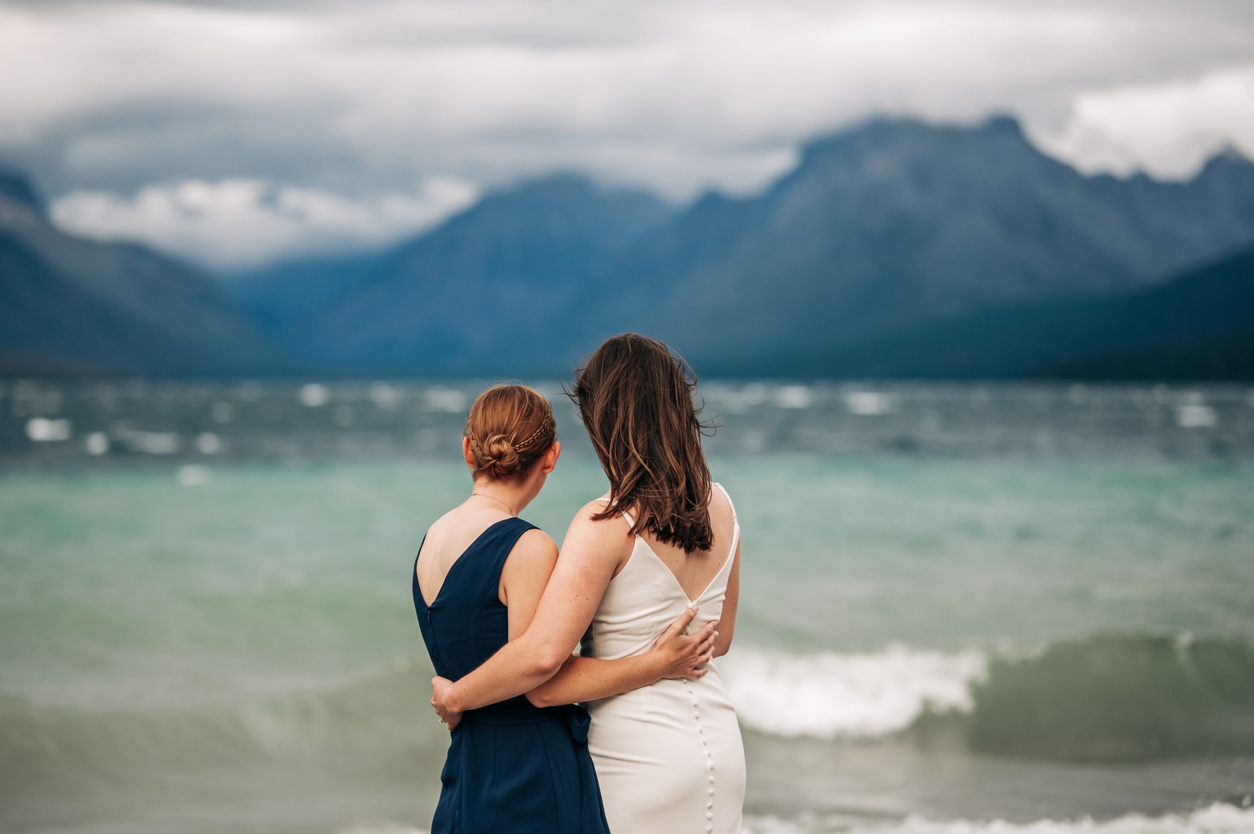 Two women embrace while gazing at a stunning mountain lake under dramatic cloudy skies.