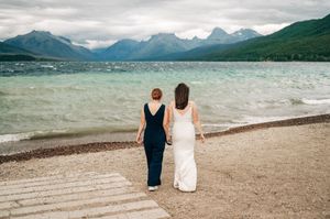 Two brides walk hand in hand along a scenic lakeside beach with turquoise water and mountain backdrop.