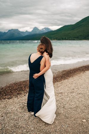 Two brides embrace on a rocky lakeshore with turquoise water and misty mountains behind them.