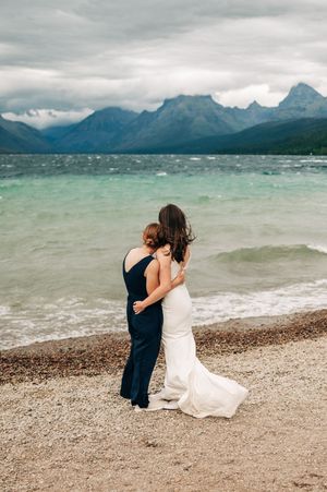 Two brides embrace on a rocky lakeshore with turquoise water and dramatic mountain peaks under cloudy skies.