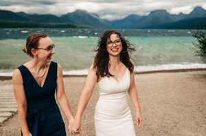 Two brides holding hands and smiling on a lakeside beach with mountain views on their wedding day.