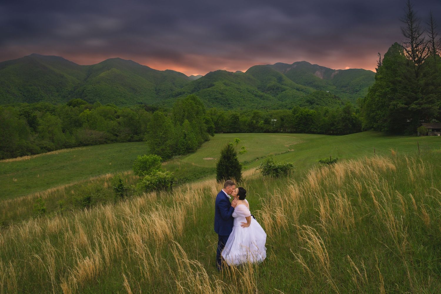 Couple embracing in lush green meadow with dramatic mountain backdrop and vibrant sunset sky.