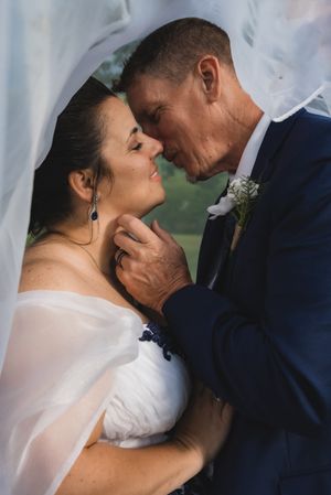 A bride and groom share a romantic moment under a veil, the groom tenderly holding her face as they lean in for a kiss.