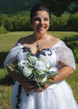 Smiling bride in white off-shoulder gown with navy lace holds white rose bouquet outdoors with green hills behind her.